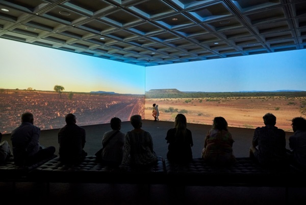 Audience watching a two-channel video of Australia's outback as the shadowed figure of a child runs in front of the projection"